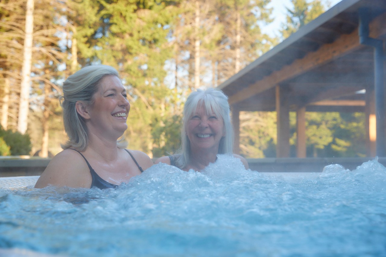two women smiling and relaxing in a bubbling hydrotherapy hot tub surrounded by forest trees