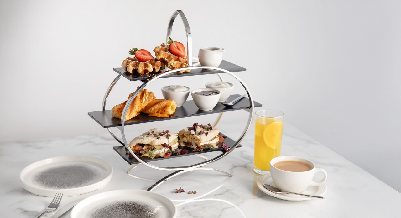 a three-tier breakfast stand featuring waffles with strawberries, pastries, small dipping sauce bowls, and sandwiches, served alongside orange juice and a cup of coffee