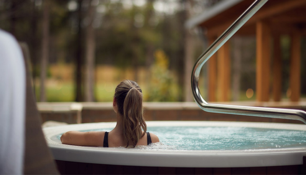 Person with a ponytail relaxes, shoulders submerged, in a bubbling round hot tub; a stainless handrail curves nearby; background shows a wooden structure and a blurred, forested outdoor setting.