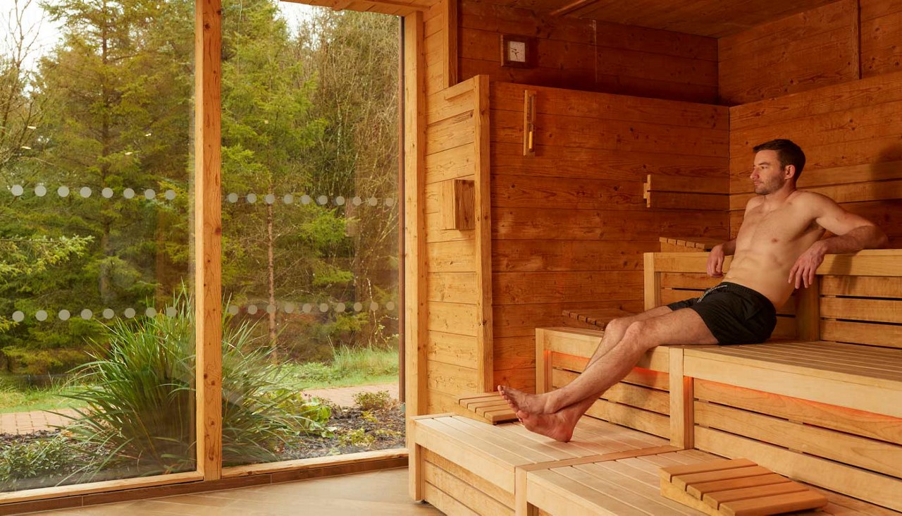 Man reclines on wooden bench, relaxing with one leg extended. Inside a timber sauna with warm lighting, next to a large window overlooking green trees and shrubs.