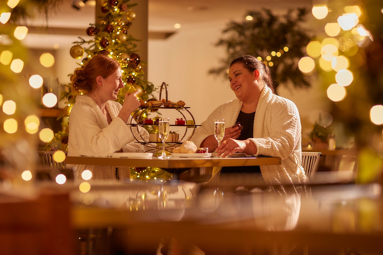 two women in spa robes sat a wooden table with christmas decorations and christmas lights surrounding them