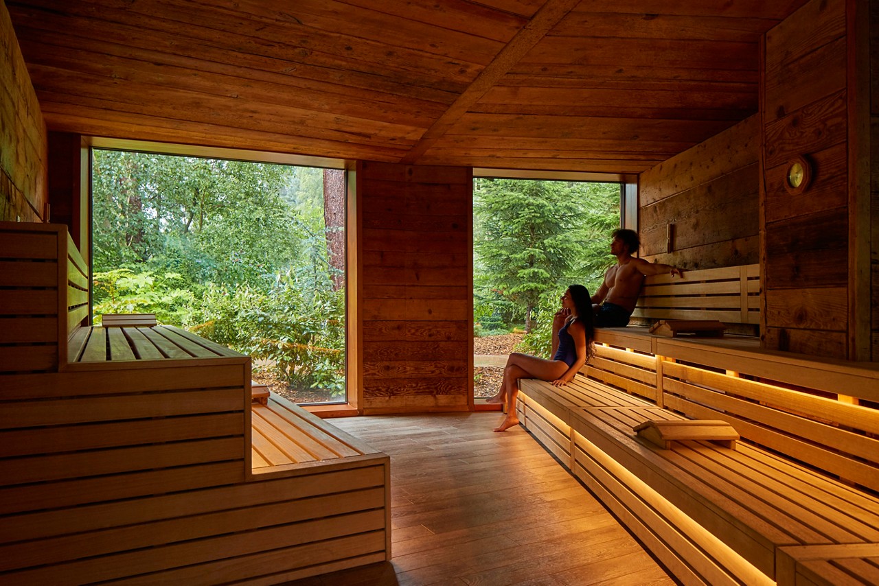 Two people relax on tiered wooden benches, sitting and leaning back, inside a warm-lit sauna; through large windows, lush green trees and garden foliage surround the quiet outdoor setting.
