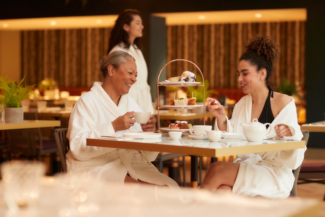 Two people in white bathrobes sip tea and choose pastries from a three-tier stand at a cafe table under warm lighting, while another robed guest stands in the background.