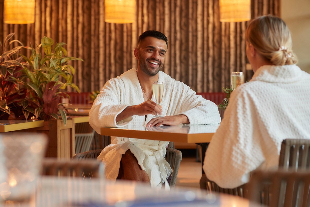 a man and women sat at a table in spa robes with glasses of prosecco