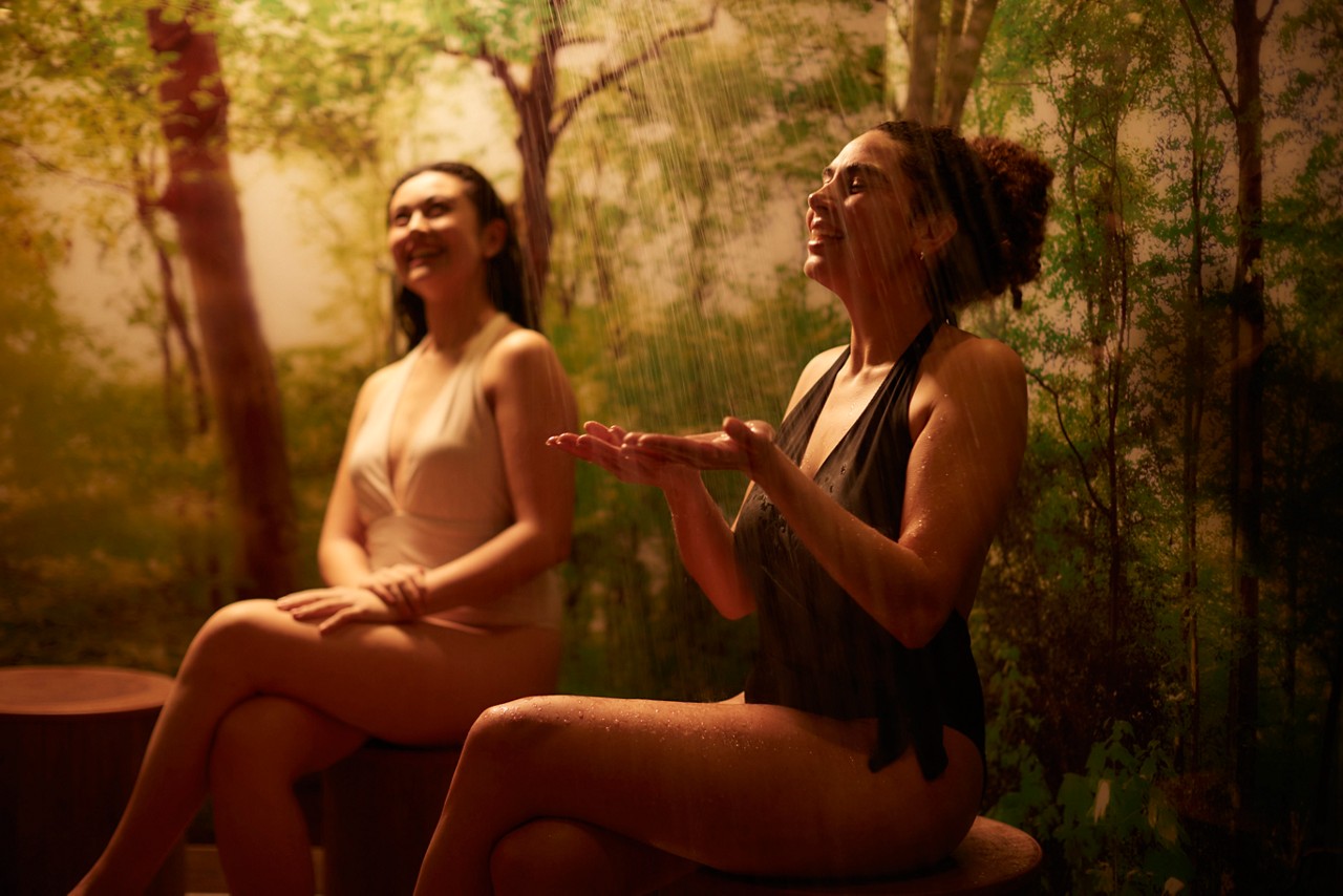 Two women in swimsuits sit on round wooden stools, catching warm shower streams and rubbing arms, in a dim, spa-like room with a forest mural background and soft golden light.
