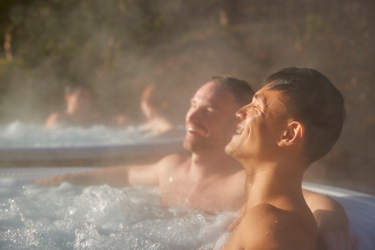 Two men soaking in an outdoor hot tub surrounded by the forest.