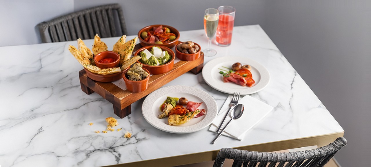 Tapas bowls present assorted appetizers—crackers, dip, olives, mozzarella with pesto, mushrooms, artichokes, cured meats—served on a wooden stand, with plated portions, cutlery, and drinks on a marble table.