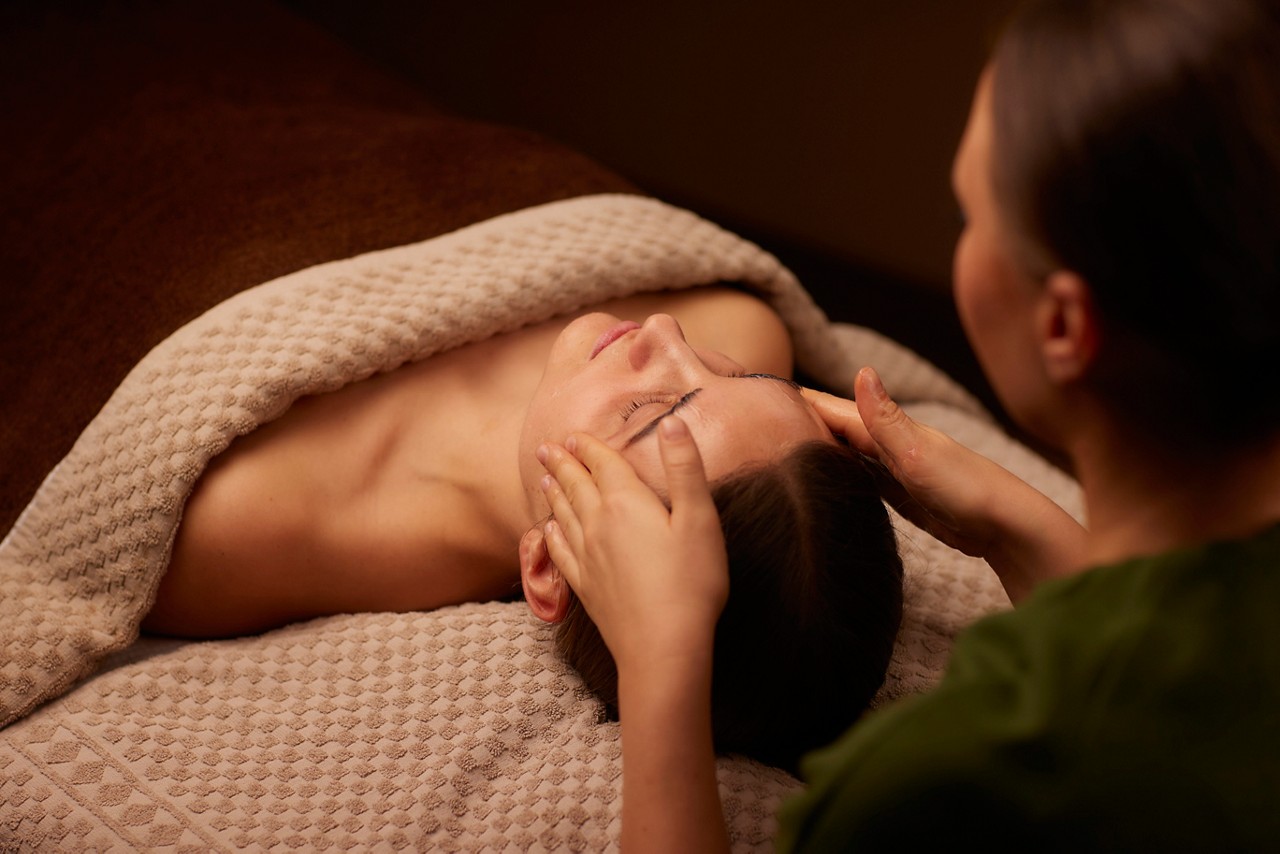 A spa therapist giving a facial treatment to client lying on a spa bed