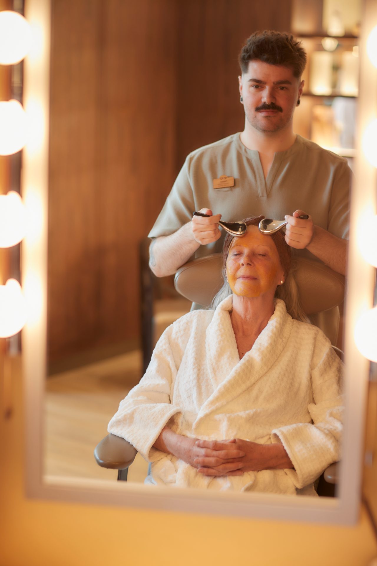 A lady sat relaxing with her eyes closed while a therapist has ice rollers on her face.