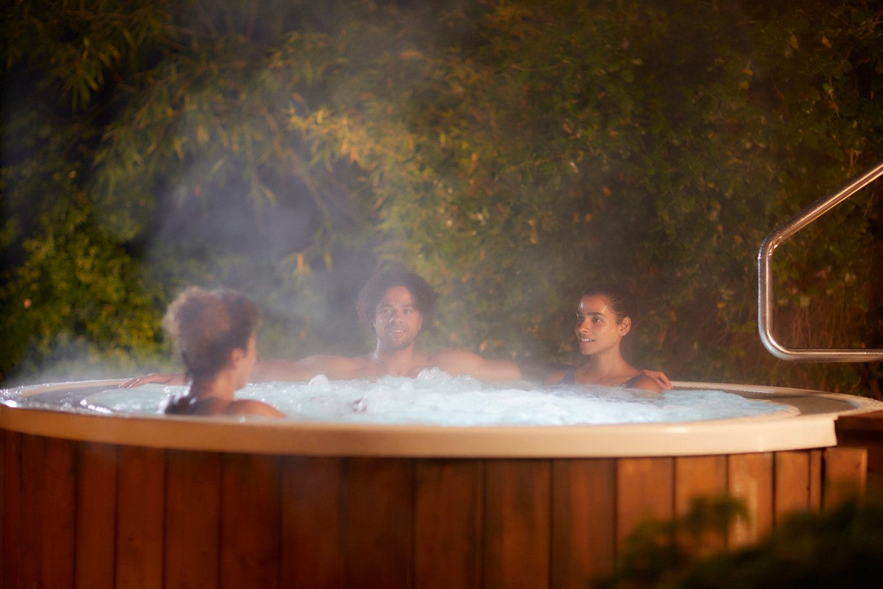 Three people relax and chat, sitting in a round, bubbling hot tub. Steam rises around them. Context: outdoor nighttime garden, leafy backdrop and metal handrail beside the wooden-sided tub.